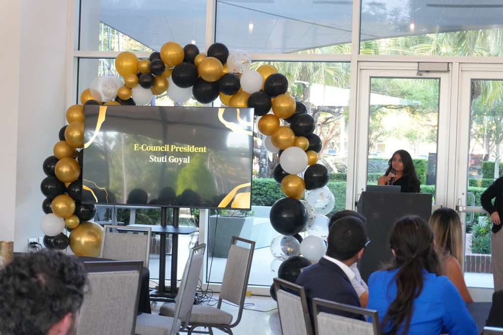 Stuti speaking at a podium beside a screen introducing her as E-Council President